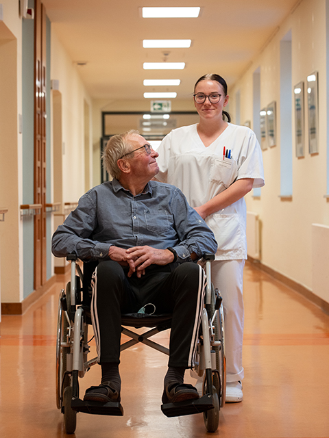 Ein Mann im Rollstuhl lächelt eine Krankenschwester an, die den Rollstuhl schiebt.